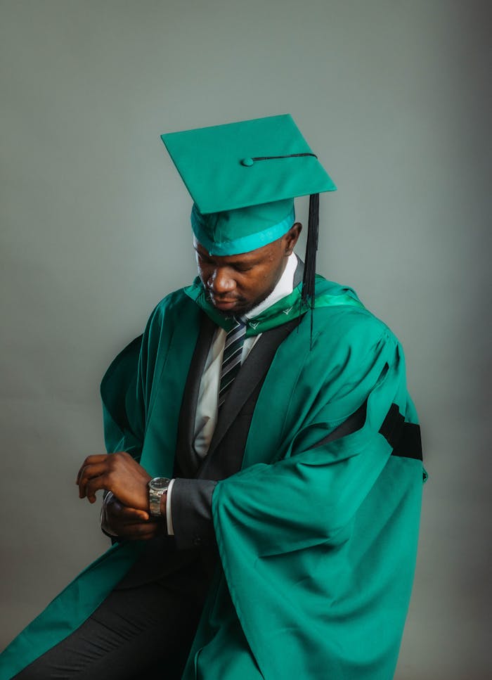 pexels-photo-30419727 Portrait of a young African graduate wearing a green cap and gown, looking at watch.