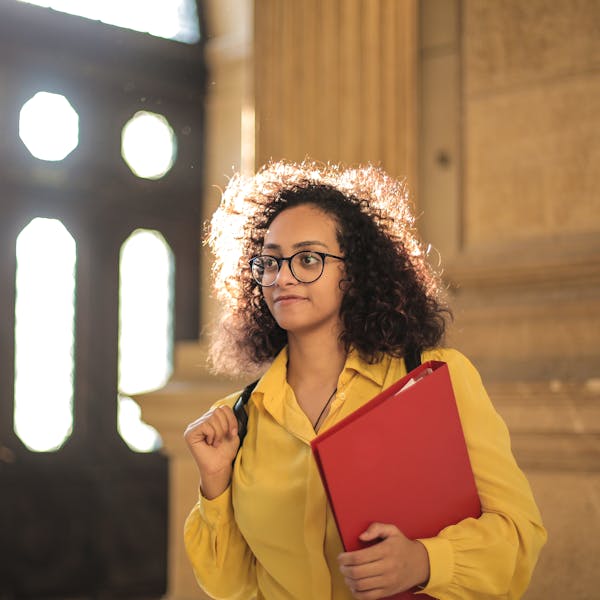 A young woman in a library carrying a red binder, bathed in sunlight.